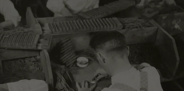 Vintage black-and-white photograph of a worker carefully hand-rolling cigars at a traditional cigar factory.