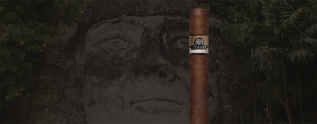 Atabey Black cigar standing upright in front of a stone-carved face and lush tropical foliage, highlighting the cigar's rich brown wrapper and detailed gold and black band.