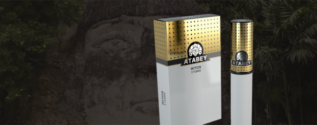Atabey Ritos cigar packaging featuring a sleek white box with metallic gold accents and three-count tube labeling, displayed beside a single matching gold and white cigar tube against a backdrop of a carved stone face and lush jungle foliage.