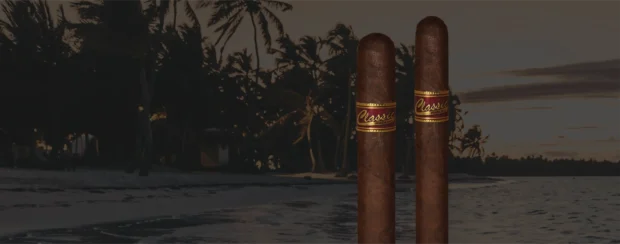 Two Classic Maduro cigars with rich dark wrappers and bold red-and-gold bands, standing on a tropical beach at sunset, capturing the essence of a full-bodied cigar experience in a relaxed coastal setting.