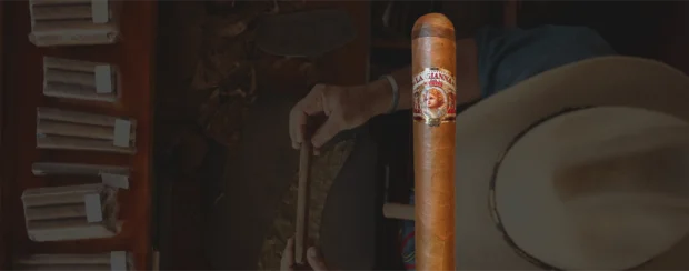 La Gianna Havana Angelic cigar with a delicate band featuring a cherub illustration, showcased against a background of a cigar artisan hand-rolling tobacco, highlighting craftsmanship and tradition.