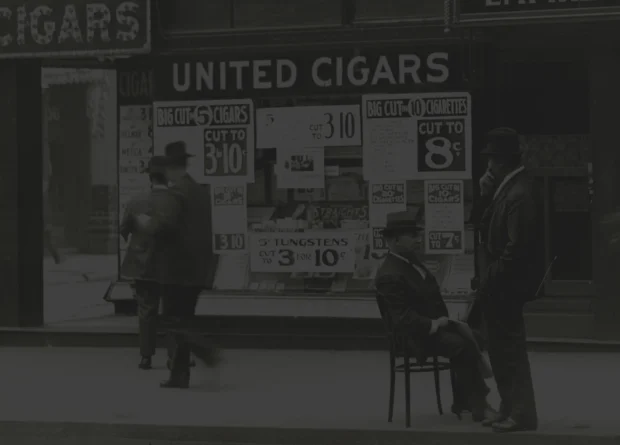Historic black-and-white photo of a United Cigars storefront featuring large promotional signs for discounted cigars and cigarettes. Several men in early 20th-century attire stand and sit on the sidewalk in front of the shop, evoking a vintage urban street scene.