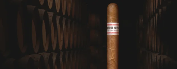 Terra Nova Dark Fired Kentucky cigar with a rustic white and red band, displayed in a dimly lit aging room lined with oak barrels, evoking bold flavor and rich, smoky tradition.