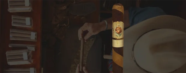 La Gianna Havana 30th Anniversary cigar with gold and cream band featuring angel artwork, displayed against a background of a cigar craftsman rolling tobacco leaves.